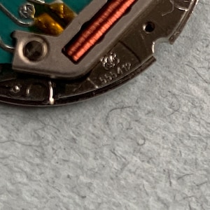 May include: Close-up of a watch movement with intricate details. The image shows a silver-colored metal component with copper-colored coils and a turquoise-colored circuit board. The number "555412" is visible on the metal.