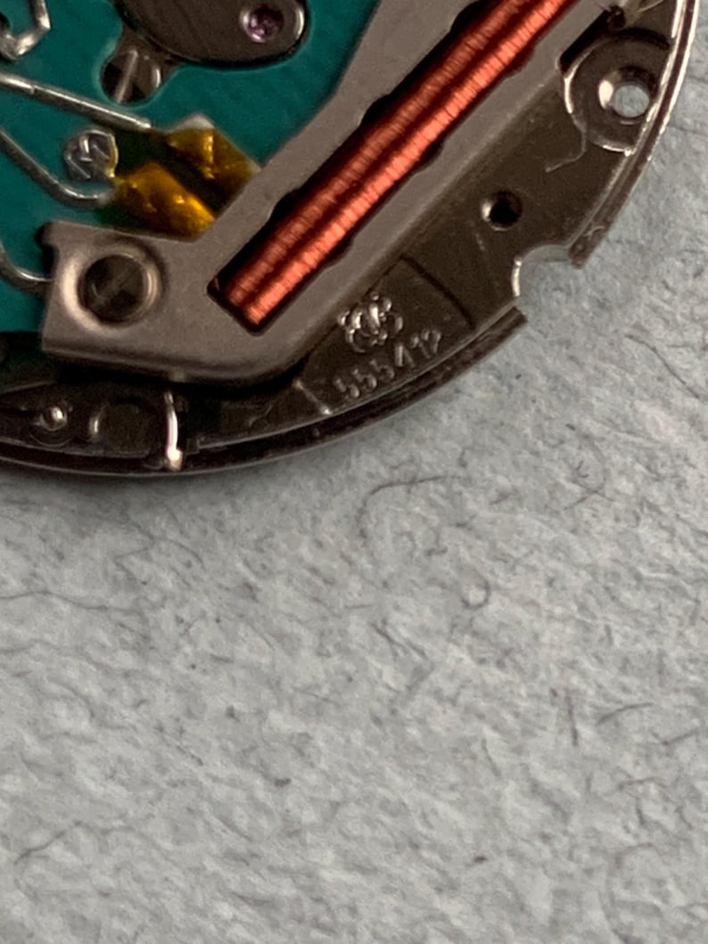 May include: Close-up of a watch movement with intricate details. The image shows a silver-colored metal component with copper-colored coils and a turquoise-colored circuit board. The number "555412" is visible on the metal.