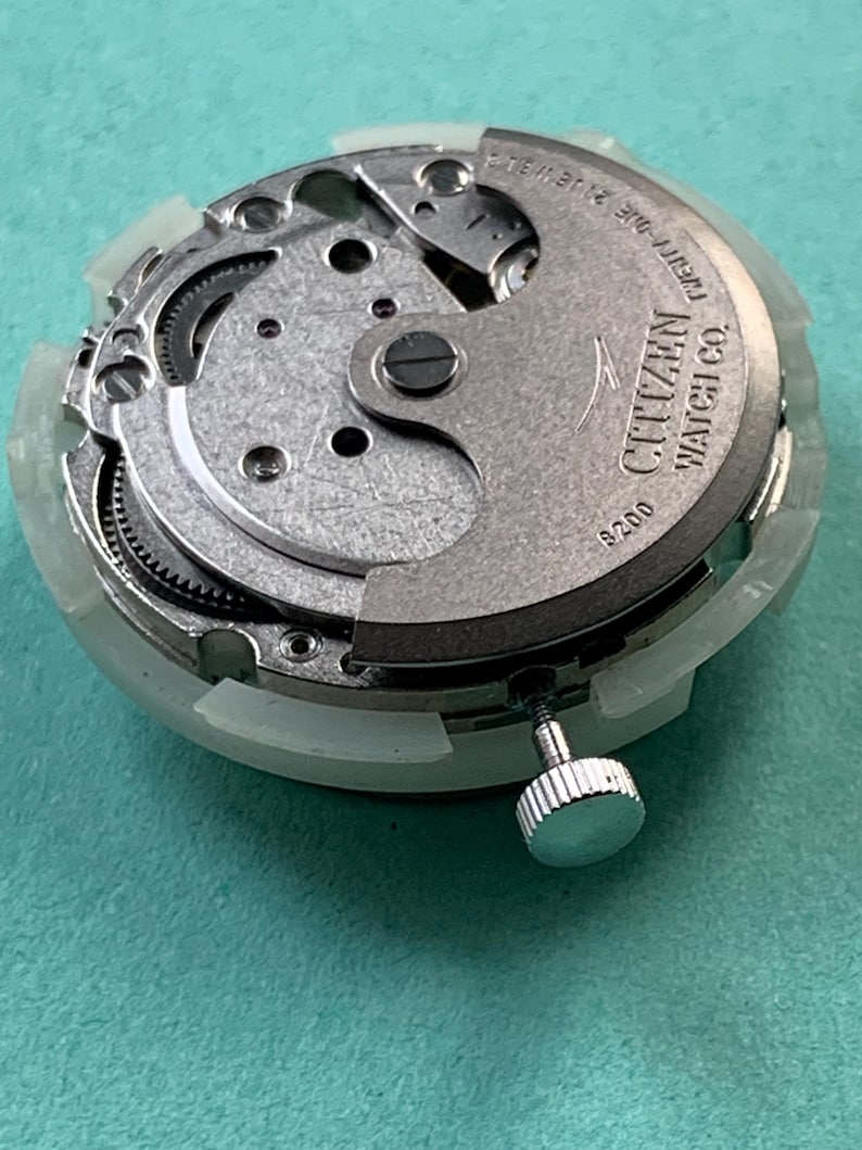 May include: A Citizen watch movement with visible gears, screws, and a crown. The metal components are silver and gray, with the brand name "Citizen Watch Co." and "Twenty-One 21 Jewels" printed on the mechanism. The movement is encased in a white plastic ring.