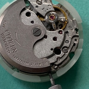 May include: Close-up of a Citizen watch movement with intricate gears and components. The text "Citizen Watch Co." and "Twenty-One 21 Jewels" is visible. The movement is set in a white plastic casing, with a silver crown.