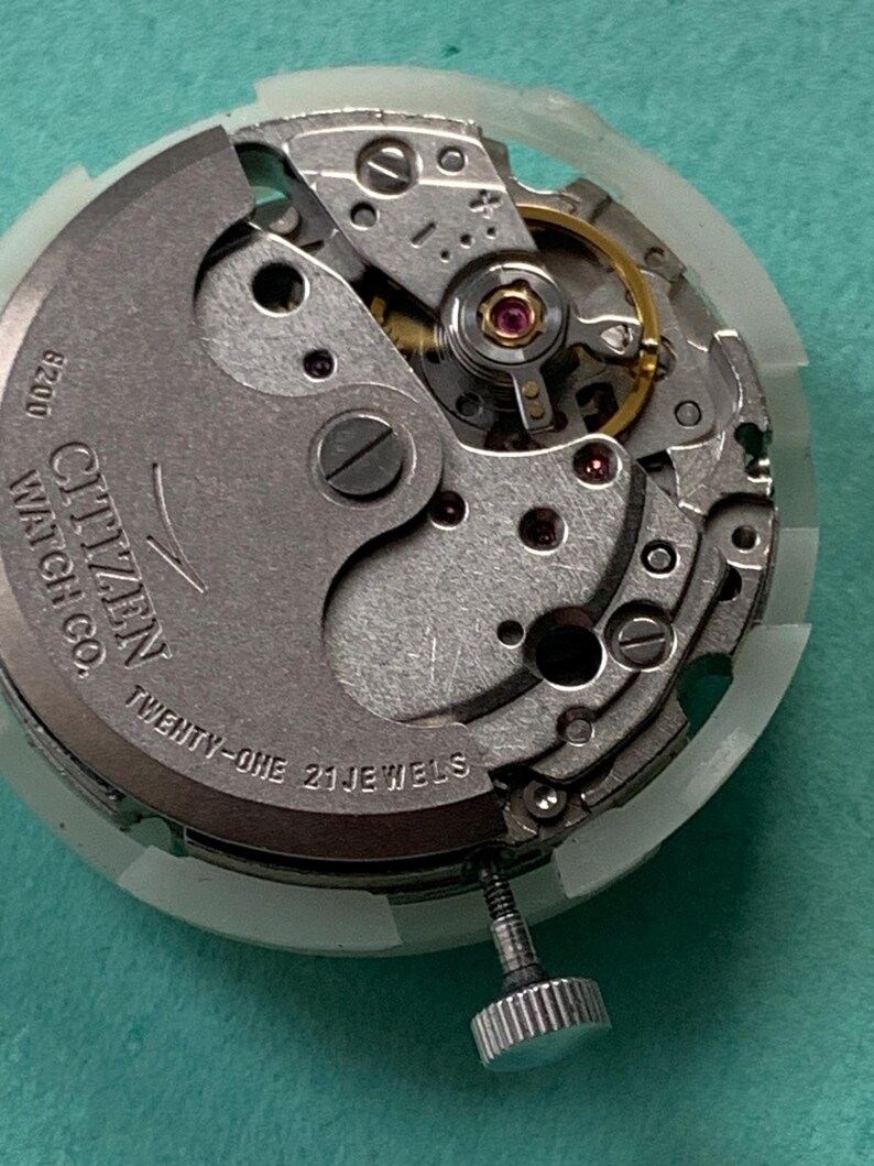 May include: Close-up of a Citizen watch movement with intricate gears and components. The text "Citizen Watch Co." and "Twenty-One 21 Jewels" is visible. The movement is set in a white plastic casing, with a silver crown.