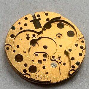 May include: Close-up of a gold-colored watch movement with intricate gears and circular cutouts. The number "2571" is visible. The watch mechanism has a complex design with multiple layers and small holes.