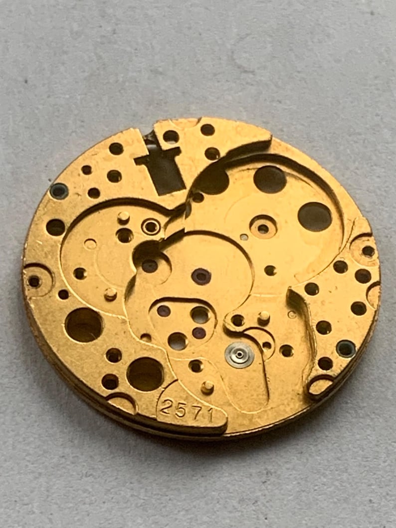 May include: Close-up of a gold-colored watch movement with intricate gears and circular cutouts. The number "2571" is visible. The watch mechanism has a complex design with multiple layers and small holes.