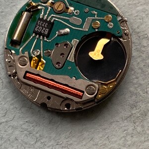 May include: Close-up of a watch movement with a green circuit board. The board features various electronic components, including a battery, integrated circuits, and a copper coil. The components are mounted on a metal frame.