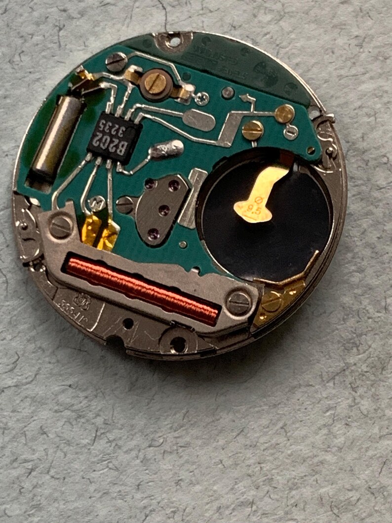 May include: Close-up of a watch movement with a green circuit board. The board features various electronic components, including a battery, integrated circuits, and a copper coil. The components are mounted on a metal frame.
