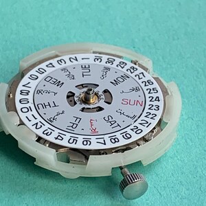 May include: Close-up of a watch movement with a white dial and Arabic numerals. The dial displays the days of the week in English and Arabic, along with the date. A silver crown is visible on the side. The background is a solid teal color.