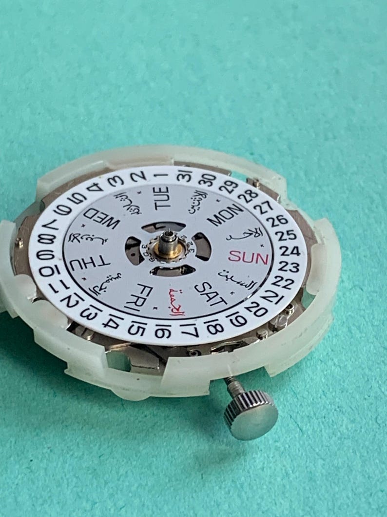 May include: Close-up of a watch movement with a white dial and Arabic numerals. The dial displays the days of the week in English and Arabic, along with the date. A silver crown is visible on the side. The background is a solid teal color.