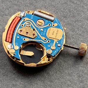 May include: A close-up of a watch movement with a blue circuit board, gold-colored components, and a winding stem. The movement has the text "ZENITH" and "SWISS V8" printed on it. The watch movement has 7 jewels.