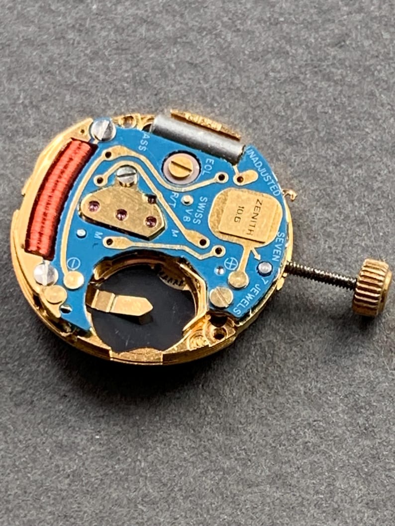 May include: A close-up of a watch movement with a blue circuit board, gold-colored components, and a winding stem. The movement has the text "ZENITH" and "SWISS V8" printed on it. The watch movement has 7 jewels.