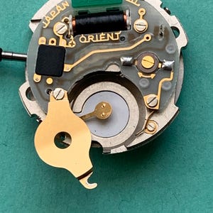 May include: Close-up of a watch movement with the word "ORIENT" printed on the circuit board. The movement has gold-colored components, including a balance wheel and screws. The text "JAPAN" is also visible.