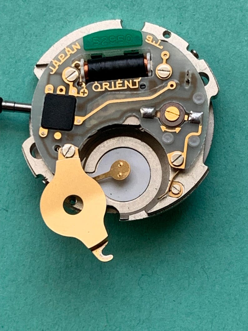 May include: Close-up of a watch movement with the word "ORIENT" printed on the circuit board. The movement has gold-colored components, including a balance wheel and screws. The text "JAPAN" is also visible.