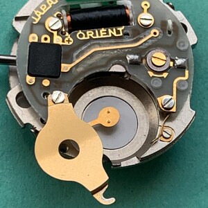 May include: Close-up of a watch movement with the brand name "ORIENT" in gold lettering. The movement has various components, including a black coil, a green piece with the number "52950", and the word "JAPAN".
