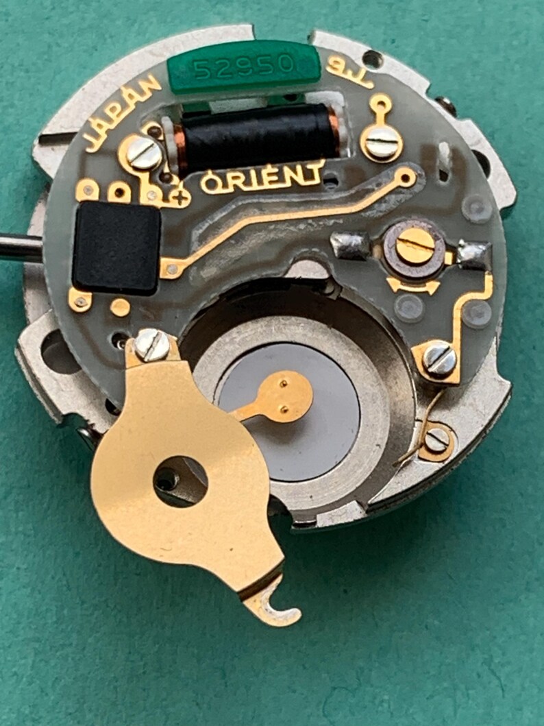 May include: Close-up of a watch movement with the brand name "ORIENT" in gold lettering. The movement has various components, including a black coil, a green piece with the number "52950", and the word "JAPAN".