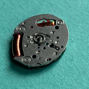 May include: Close-up of a watch movement with a silver-colored metal base. The movement features various small holes, gears, and a copper-colored coil. The background is a solid teal color.