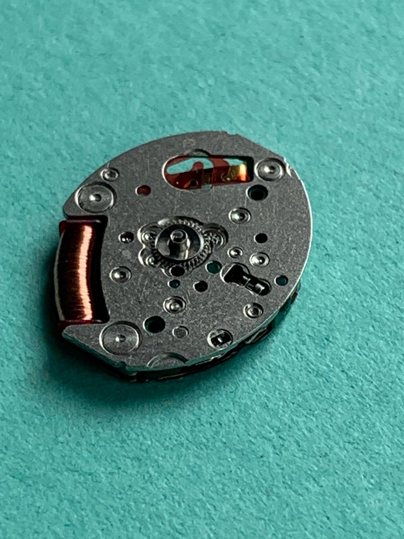 May include: Close-up of a watch movement with a silver-colored metal base. The movement features various small holes, gears, and a copper-colored coil. The background is a solid teal color.