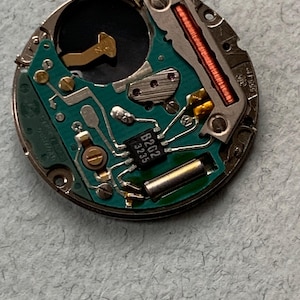 May include: Close-up of a watch movement with a green circuit board, metallic components, and a copper coil. The board has the text "B202 3235". The watch mechanism is circular and has a silver metal casing.