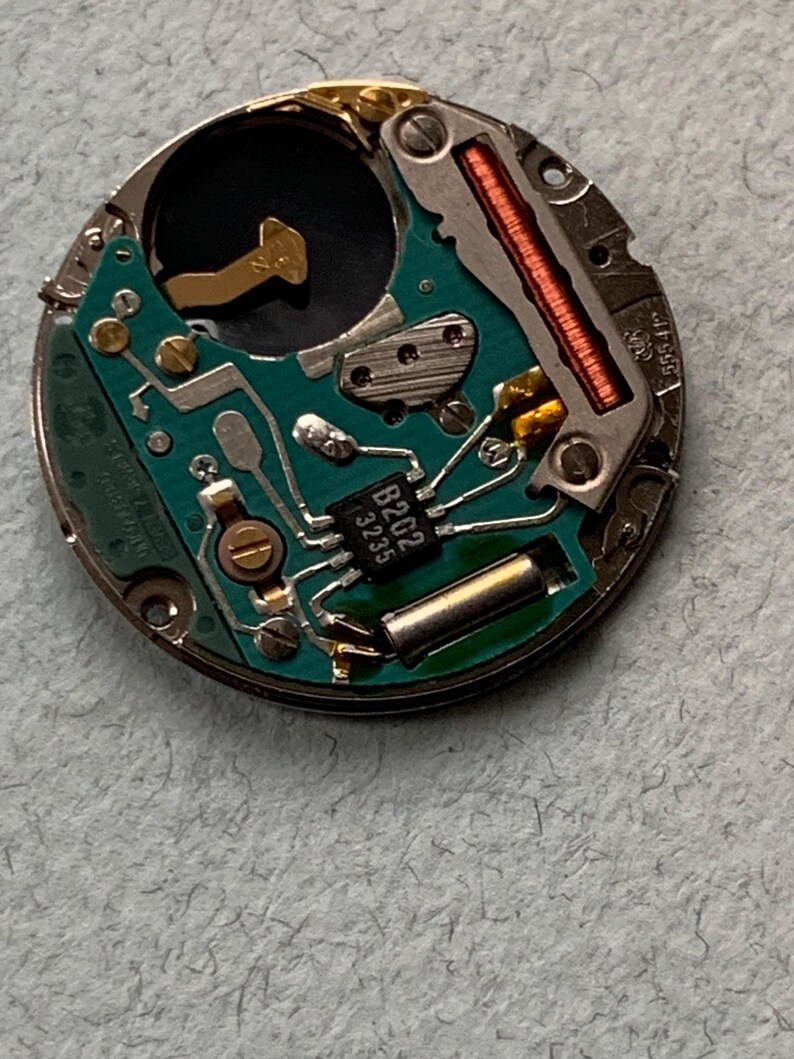 May include: Close-up of a watch movement with a green circuit board, metallic components, and a copper coil. The board has the text "B202 3235". The watch mechanism is circular and has a silver metal casing.