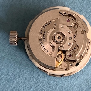 May include: A Citizen watch movement with 21 jewels, made in Japan. The silver-colored mechanism features intricate gears, red jewels, and a winding stem. The watch movement is set against a light blue background.