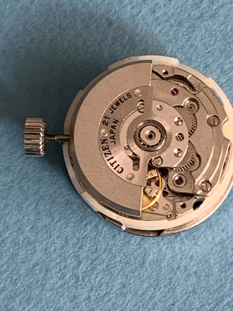 May include: A Citizen watch movement with 21 jewels, made in Japan. The silver-colored mechanism features intricate gears, red jewels, and a winding stem. The watch movement is set against a light blue background.