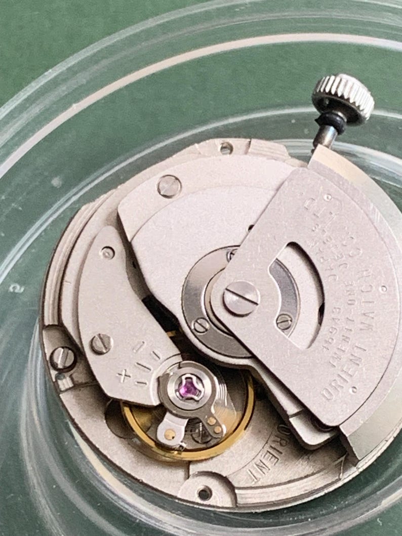May include: Close-up of a silver-toned Orient Watch movement. The watch mechanism features various gears, screws, and a crown. The text "ORIENT WATCH" is visible on the movement. The watch movement is set in a clear, circular container.