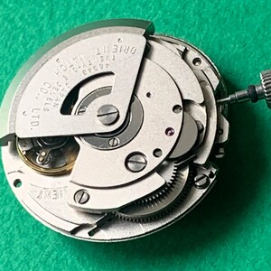 May include: A close-up of a silver Orient watch movement with intricate gears, jewels, and the brand name visible. The watch mechanism is set against a green background, showcasing the internal components of the timepiece.