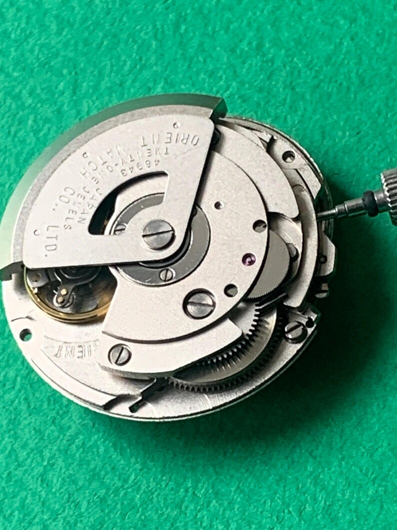 May include: A close-up of a silver Orient watch movement with intricate gears, jewels, and the brand name visible. The watch mechanism is set against a green background, showcasing the internal components of the timepiece.