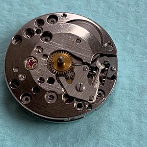 May include: A silver watch movement with a gold gear and a pink jewel. The movement is disassembled and shows the inner workings of the watch.