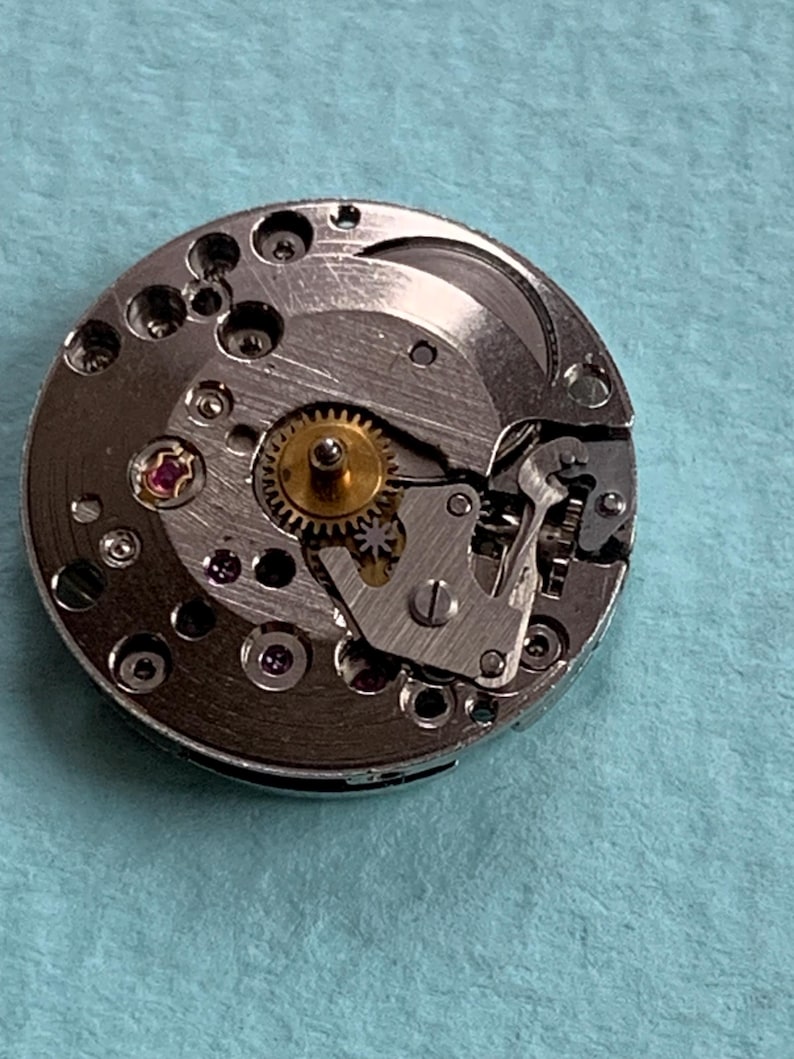 May include: A silver watch movement with a gold gear and a pink jewel. The movement is disassembled and shows the inner workings of the watch.
