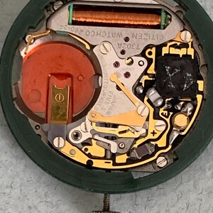 May include: Close-up of a Citizen watch movement, showcasing intricate internal components. The watch mechanism features a copper-colored coil, a red battery, and various gold-toned gears and levers. The green outer casing and silver adjustment screw are also visible.