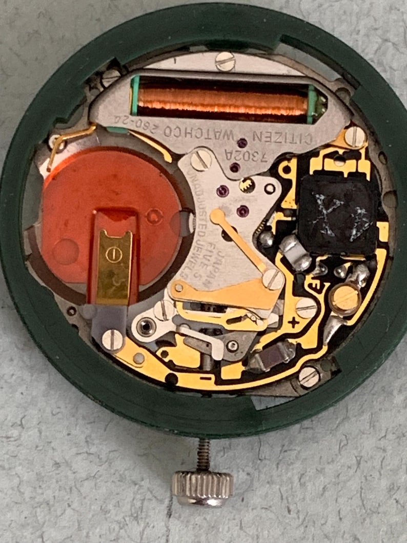 May include: Close-up of a Citizen watch movement, showcasing intricate internal components. The watch mechanism features a copper-colored coil, a red battery, and various gold-toned gears and levers. The green outer casing and silver adjustment screw are also visible.