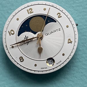Op de afbeelding: Een witte quartz wijzerplaat met gouden cijfers en wijzers. De wijzerplaat heeft een maanfase-display en het woord "Royal". De datum is ingesteld op 19. "Swiss Made" staat onderaan gedrukt.