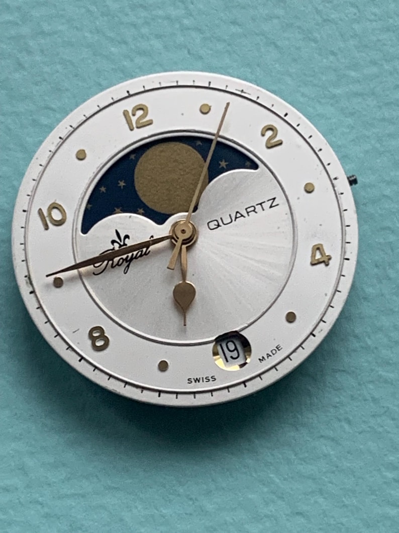 Op de afbeelding: Een witte quartz wijzerplaat met gouden cijfers en wijzers. De wijzerplaat heeft een maanfase-display en het woord "Royal". De datum is ingesteld op 19. "Swiss Made" staat onderaan gedrukt.