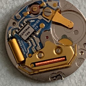Może przedstawiać: Zbliżenie mechanizmu zegarka Certina 729 Swiss V8. Mechanizm zegarka posiada złote elementy, niebieską płytkę obwodu drukowanego oraz tekst "CERTINA 729" i "SWISS V8". Mechanizm zegarka jest w kolorze srebrnym.
