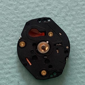 May include: A black watch movement featuring gold screws and a small, silver gear at the centre. A red-orange component is visible. The watch mechanism is set against a light blue background.