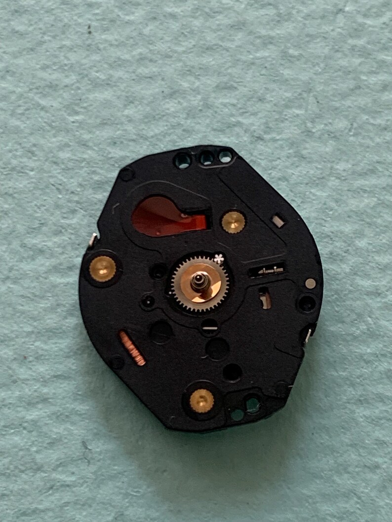 May include: A black watch movement featuring gold screws and a small, silver gear at the centre. A red-orange component is visible. The watch mechanism is set against a light blue background.