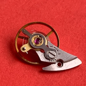 May include: Close-up of a watch escapement mechanism. The mechanism features a gold-coloured balance wheel, a silver-coloured lever, and a small, round, purple jewel. The background is a solid red colour.