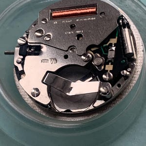 Op de afbeelding: Close-up van een uurwerk met diverse componenten. Het uurwerk heeft een zilverkleurige metalen basis met schroeven, een batterij en een koperkleurige spoel. Het uurwerk bevindt zich in een transparante, ronde container.