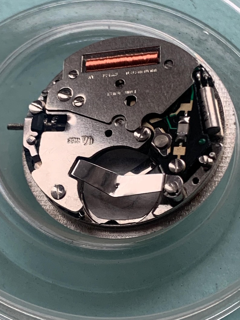 Op de afbeelding: Close-up van een uurwerk met diverse componenten. Het uurwerk heeft een zilverkleurige metalen basis met schroeven, een batterij en een koperkleurige spoel. Het uurwerk bevindt zich in een transparante, ronde container.