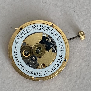 May include: A gold-toned watch movement with a white date dial featuring black numerals from 1 to 31. The intricate mechanism includes gears and a screw-down crown. The watch part is designed for repair or restoration.