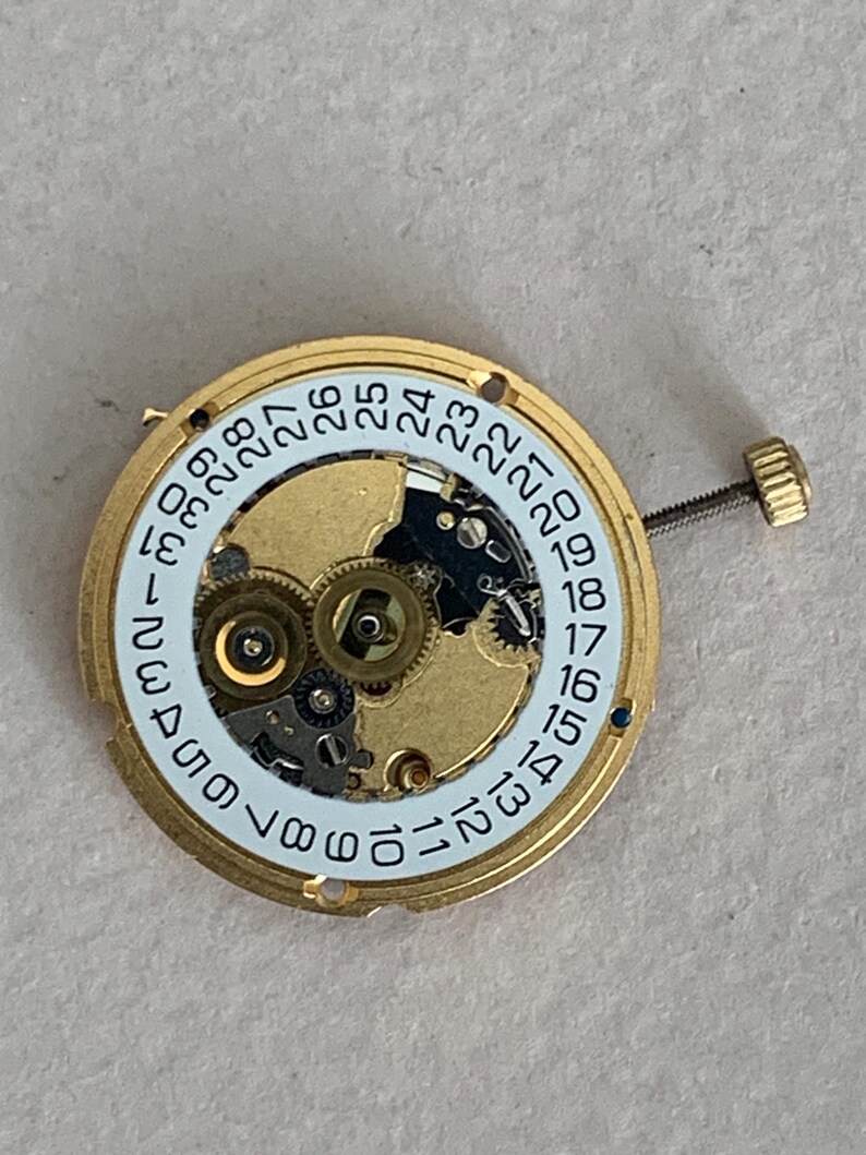 May include: A gold-toned watch movement with a white date dial featuring black numerals from 1 to 31. The intricate mechanism includes gears and a screw-down crown. The watch part is designed for repair or restoration.