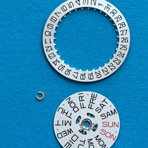 May include: Watch parts on a blue background. The image shows a silver-colored date wheel with numbers 1-31, a day wheel with days of the week, and a small gold-colored clip. The day wheel has the word "SUN" in red.