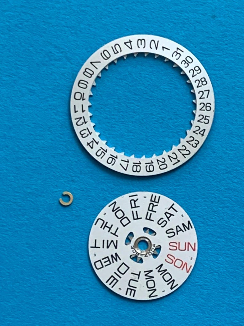 May include: Watch parts on a blue background. The image shows a silver-colored date wheel with numbers 1-31, a day wheel with days of the week, and a small gold-colored clip. The day wheel has the word "SUN" in red.