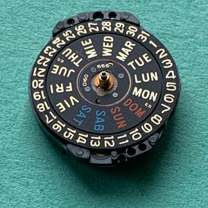 May include: A black watch calendar dial with gold-colored numbers and day abbreviations. The dial displays the days of the week in multiple languages and numbers from 1 to 31. The center has a small gold-colored post.