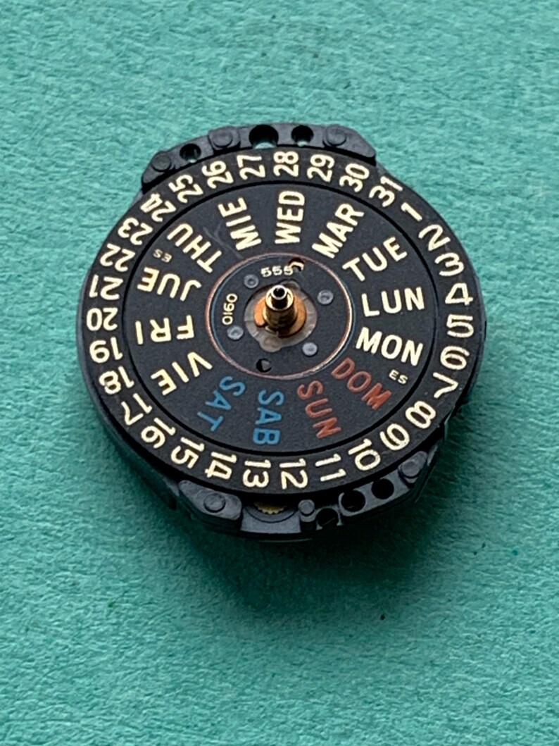 May include: A black watch calendar dial with gold-colored numbers and day abbreviations. The dial displays the days of the week in multiple languages and numbers from 1 to 31. The center has a small gold-colored post.