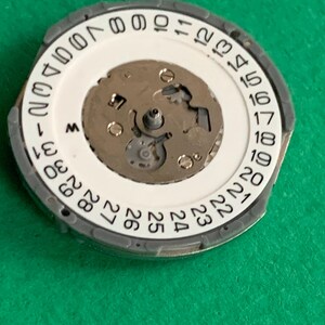 May include: A watch movement component with a white dial featuring black numerals from 1 to 31, indicating the days of the month. The component has a gray outer ring and a central mechanism with small gears and screws.