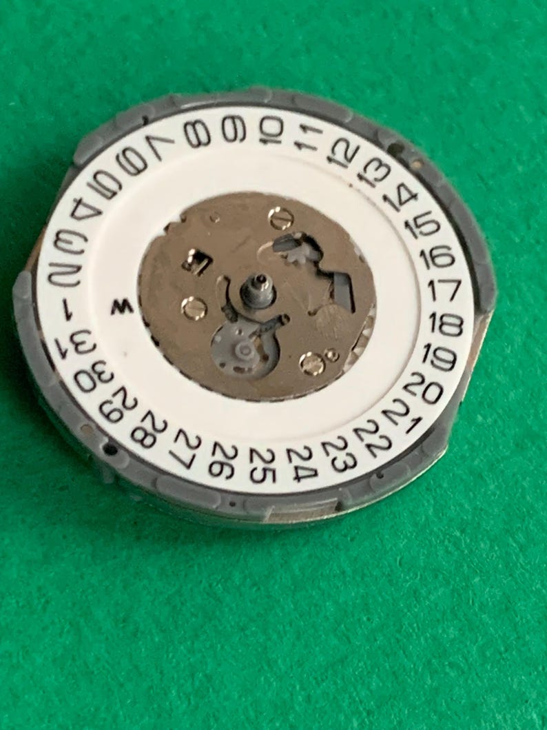 May include: A watch movement component with a white dial featuring black numerals from 1 to 31, indicating the days of the month. The component has a gray outer ring and a central mechanism with small gears and screws.