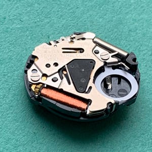 May include: Close-up of a watch movement with the text "NO JEWELS SEIKO TIME CORP 7N83D". The movement features metallic components, a copper coil, and a black plastic base, set against a teal background.