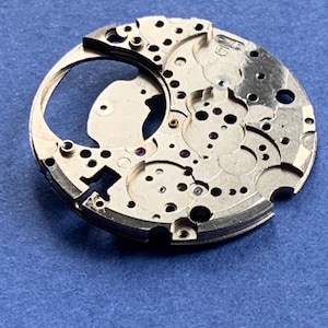 Puede incluir: Primer plano de un componente plateado de movimiento de reloj con intrincados engranajes, agujeros y peque&ntilde;as joyas de rub&iacute;. La pieza circular tiene un dise&ntilde;o complejo, mostrando ingenier&iacute;a de precisi&oacute;n. El fondo es azul liso.