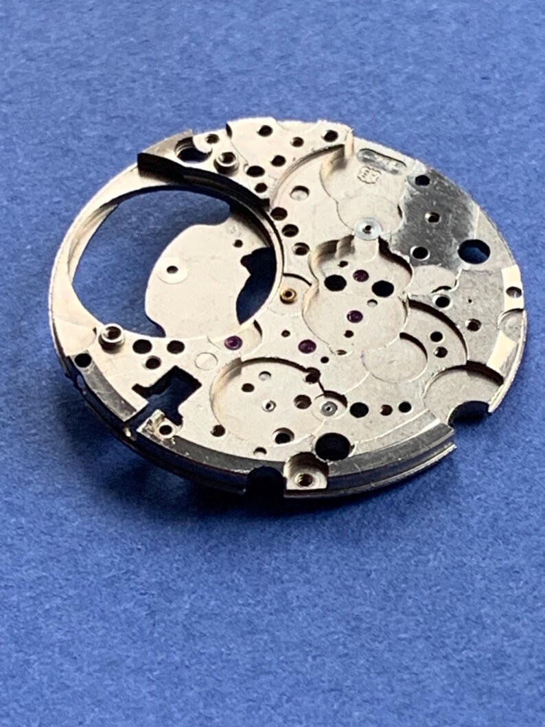 Puede incluir: Primer plano de un componente plateado de movimiento de reloj con intrincados engranajes, agujeros y peque&ntilde;as joyas de rub&iacute;. La pieza circular tiene un dise&ntilde;o complejo, mostrando ingenier&iacute;a de precisi&oacute;n. El fondo es azul liso.
