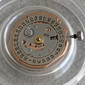 Puede incluir: Primer plano de un movimiento de reloj con una esfera plateada y dorada. La esfera presenta n&uacute;meros negros que indican la fecha y la hora. Se puede ver una peque&ntilde;a corona y otros componentes mec&aacute;nicos. El movimiento est&aacute; encerrado en un soporte de pl&aacute;stico transparente.